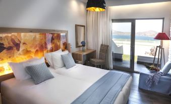 a hotel room with a large bed , white walls , and a balcony overlooking the ocean at Elounda Orama