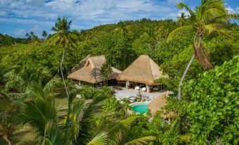 a tropical resort with thatched - roof huts , surrounded by lush greenery and clear blue skies , including a pool at North Island, a Luxury Collection Resort, Seychelles