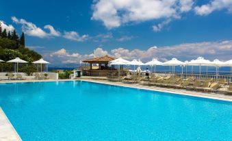 a large outdoor swimming pool surrounded by lounge chairs and umbrellas , with a view of the ocean in the background at Louis Ionian Sun