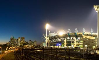 a stadium with a lit - up sign and skyline in the background , as well as train tracks leading to the entrance at Punthill Ivanhoe