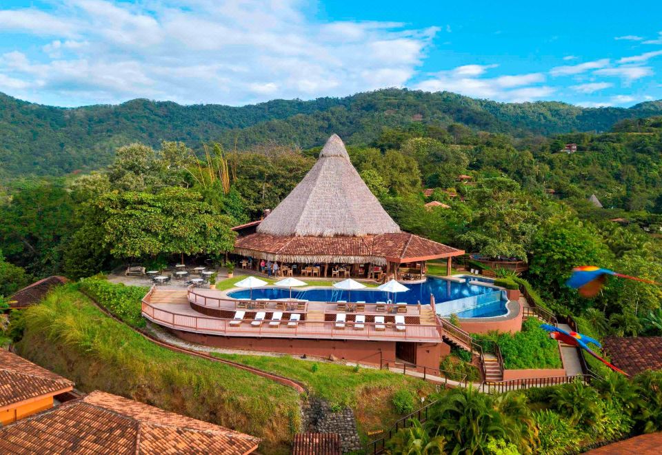 a large , thatched - roof building with a pool and lounge chairs is surrounded by lush greenery and mountains at Punta Islita, Autograph Collection