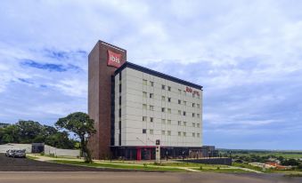 a large , modern hotel building with a red sign and brown brick exterior , surrounded by trees and grass , under a cloudy sky at Ibis Guaiba