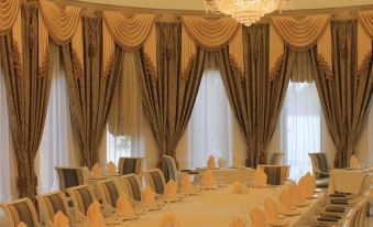 a long dining table with gold napkins and a chandelier in the center is set up in a large , ornate room with brown curtains at Druzhba