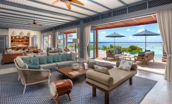 a spacious living room with a couch , chairs , and tables , overlooking a pool and the ocean at Raffles Maldives Meradhoo Resort