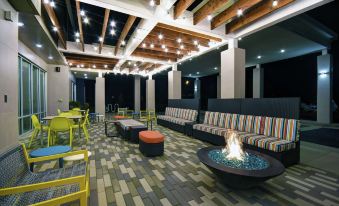 a modern lounge area with colorful chairs , a fire pit , and various seating arrangements under wooden beams at Home2 Suites by Hilton Lawrenceville Atlanta Sugarloaf