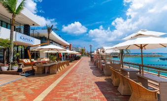 a brick walkway with rows of tables and chairs , umbrellas , and palm trees along the waterfront at Sunset Beach Hotel