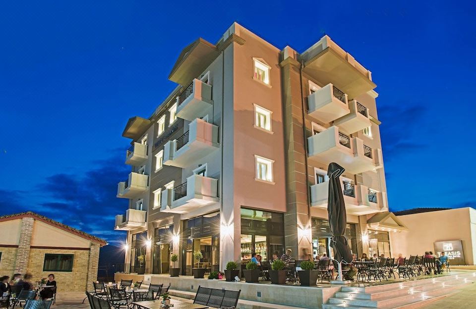 a tall , multi - story building with balconies and a fountain in front of it , surrounded by trees and other buildings at Theasis Hotel Paramythia