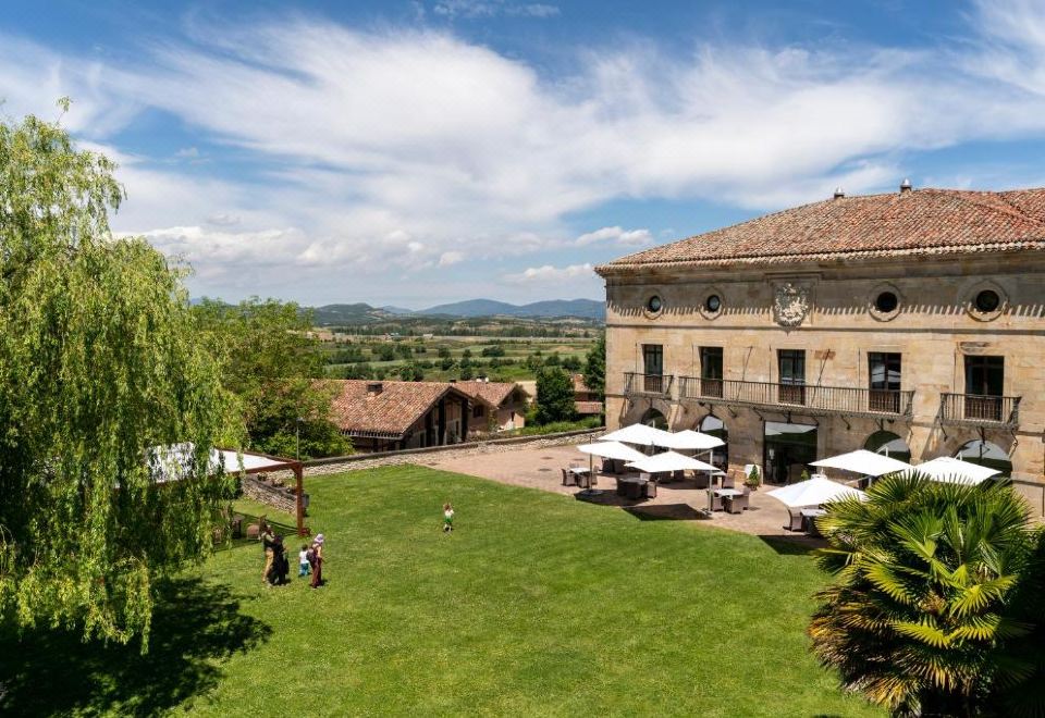 a large building with a red roof is surrounded by green grass and umbrellas , as well as people walking around at Parador de Argomaniz