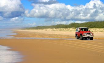 a red jeep is driving on a sandy beach , surrounded by green trees and blue sky at Noosa North Shore Retreat