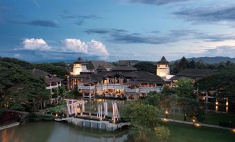 a picturesque scene of a large building surrounded by trees and a lake , illuminated at night at Le Meridien Chiang Rai Resort, Thailand