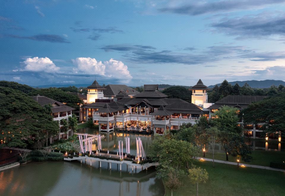 a picturesque scene of a large building surrounded by trees and a lake , illuminated at night at Le Meridien Chiang Rai Resort, Thailand