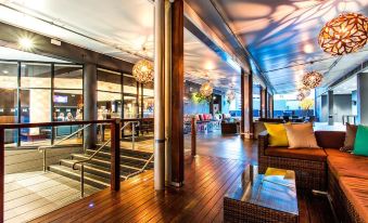 a wooden terrace with multiple lounge chairs , a table , and a reception desk , surrounded by columns and lit up by hanging lights at Hotel Grand Chancellor Brisbane