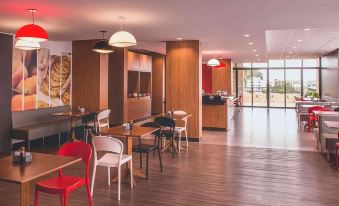 a modern restaurant with wooden floors , red chairs , and a bar area under large windows at Ibis Carlos Barbosa