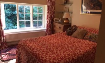 a well - lit bedroom with a large window , red patterned bedspread , and a wooden nightstand near the window at Bridge Cottage