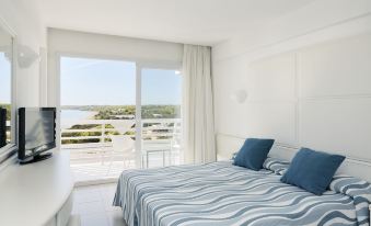 a white bedroom with a large bed , blue and white striped curtains , and a balcony overlooking the ocean at Insotel Hotel Formentera Playa