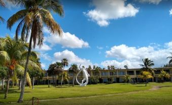 a grassy field with a large white sculpture in the middle , surrounded by palm trees and buildings at Colony