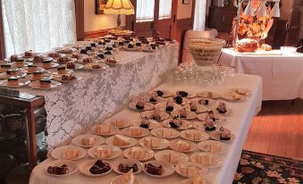 a table is set with various desserts , including cakes and pastries , on white plates and glasses , in a room with large windows at Woodbine Hotel and Restaurant