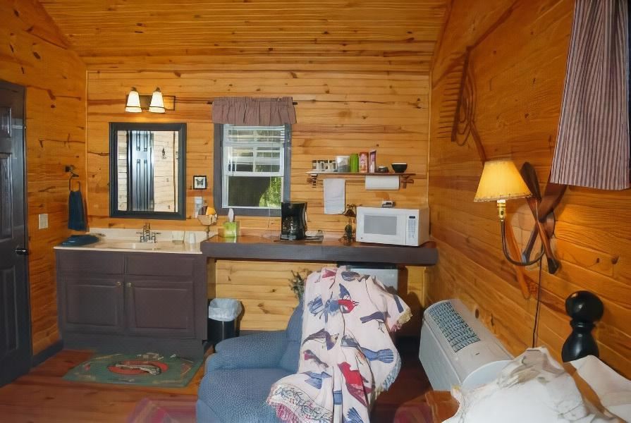 a cozy cabin interior with wooden walls , exposed beams , and a blue armchair , as well as a kitchen area with appliances and sink at River Valley Rentals