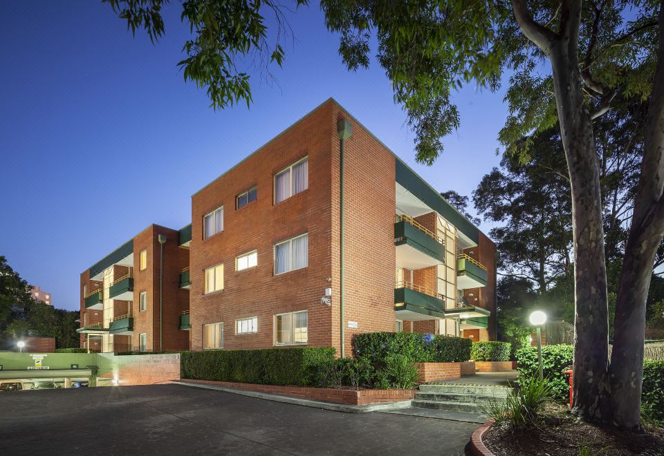 a two - story apartment building with red brick walls and green balconies , situated in a residential area at APX Parramatta