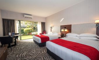 a hotel room with two beds , each bed having red and white bedding , and a sliding glass door leading to a garden at Waipuna Hotel & Conference Centre