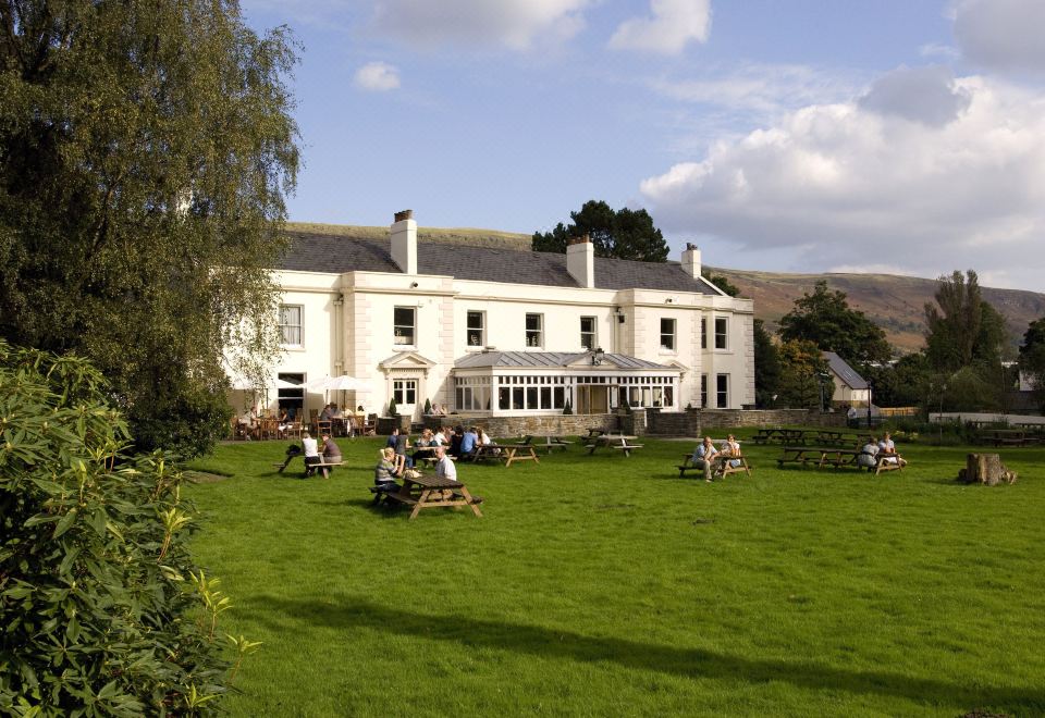 a large white building surrounded by green grass and trees , with people sitting on benches in the lawn at Premier Inn Merthyr Tydfil