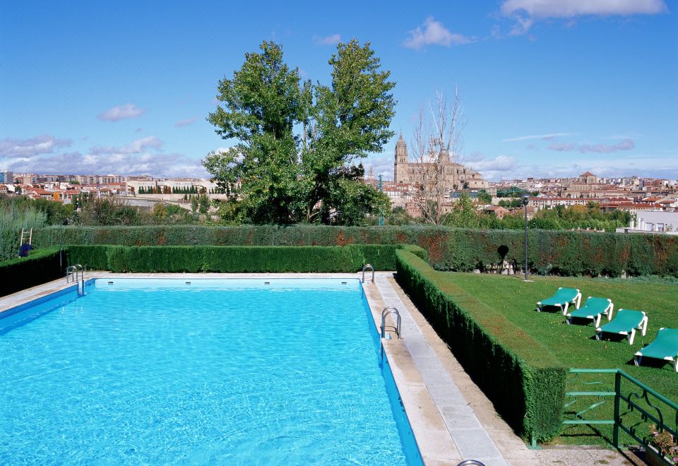 a large swimming pool with a view of the city , surrounded by green grass and trees at Parador de Salamanca