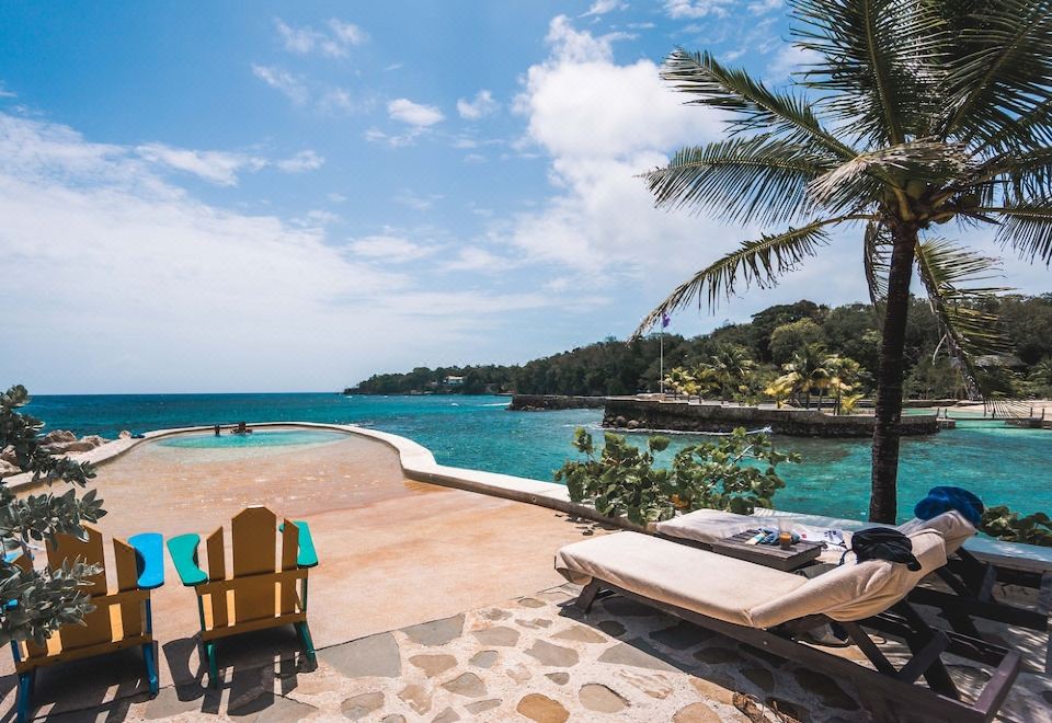 a beautiful beach scene with a large pool , lounge chairs , and a palm tree , under a blue sky dotted with clouds at GoldenEye