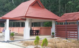 a red - roofed gazebo with a white pole , surrounded by trees and bushes , and situated in front of a building at P.K Inn
