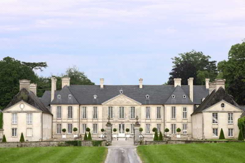 a large , white stone mansion with multiple windows and doors , surrounded by lush greenery and a clear blue sky at Chateau d'Audrieu