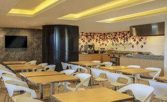 a modern cafeteria with white chairs and tables , a black vase on the counter , and yellow lights illuminating the ceiling at Fiesta Inn Express Queretaro Constituyentes