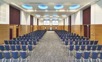 a large conference room with rows of blue chairs and a man sitting at the front at Hilton at St.George's Park, Burton Upon Trent