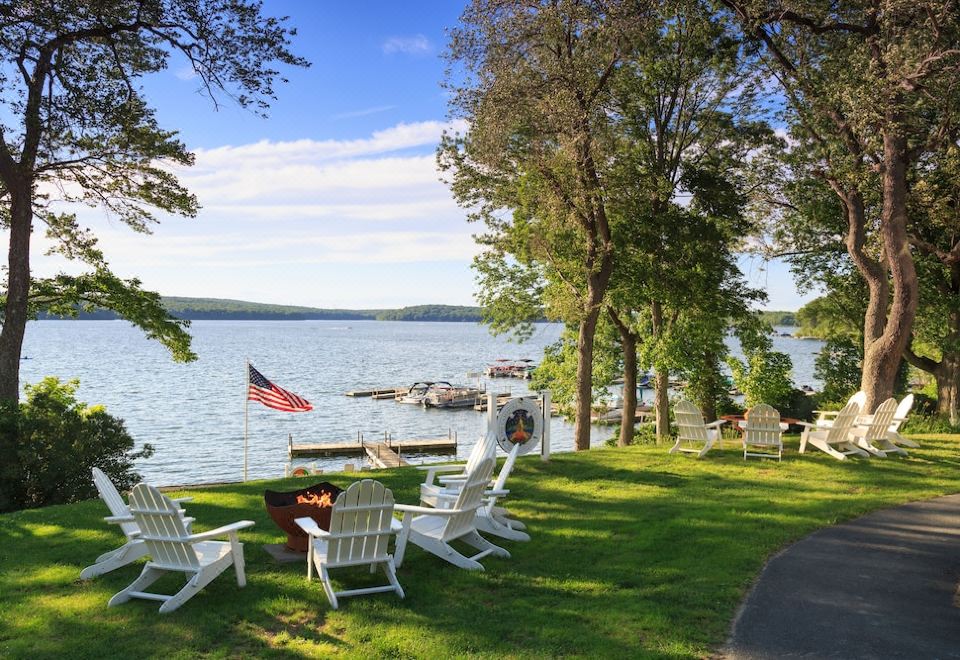 a serene outdoor setting with white lounge chairs , a bonfire , and an american flag , overlooking a calm lake surrounded by trees at Silver Birches Resort