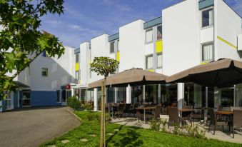 a modern building with white walls and yellow accents , surrounded by lush greenery and outdoor dining areas at Ibis Longwy Mexy