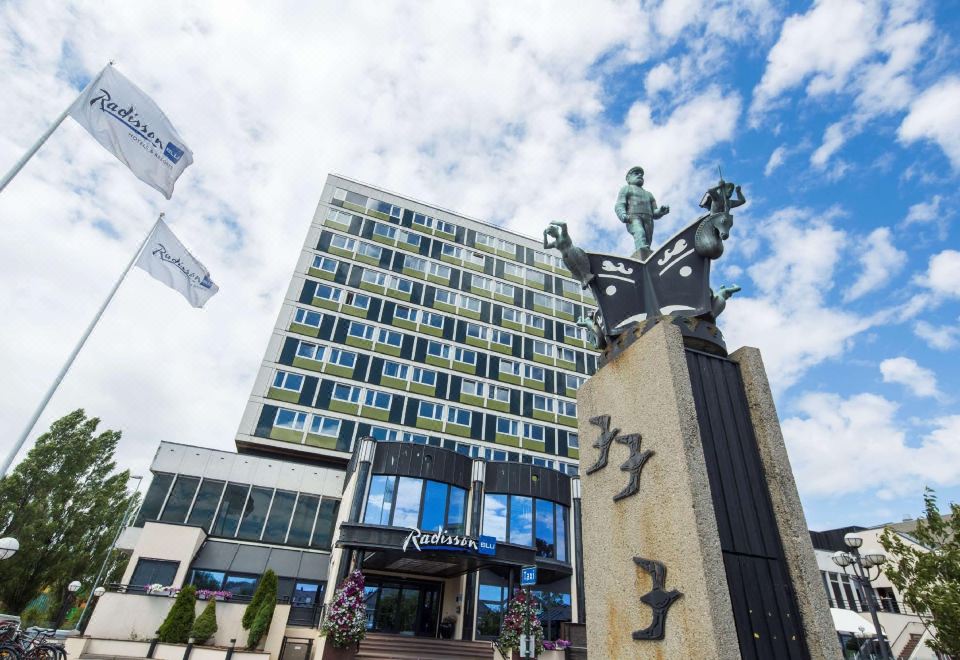 a large hotel building with a statue in front of it , surrounded by flags and people at Radisson Blu Caledonien Hotel, Kristiansand