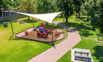 a playground with a sandy area and play structure under a large , white canopy , surrounded by green grass and trees at Adelaide Caravan Park - Aspen Holiday Parks