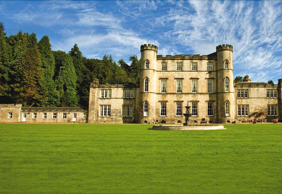 a large stone building with towers and turrets , surrounded by green grass and trees , under a clear blue sky at Melville Castle Hotel
