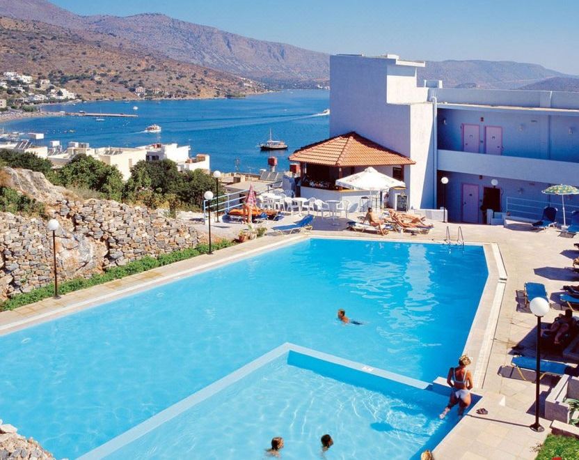a large swimming pool is surrounded by lounge chairs and a building , with a view of the ocean in the background at Elounda Orama