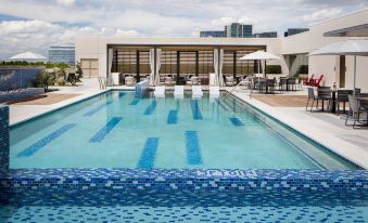 a large swimming pool with a blue tile pattern is surrounded by white chairs and tables , while in the background buildings at Renaissance Dallas at Plano Legacy West Hotel