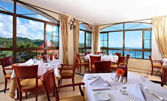 a restaurant with white tablecloths and chairs , set for dining tables , has large windows overlooking the ocean at Bahia Principe Grand Cayacoa