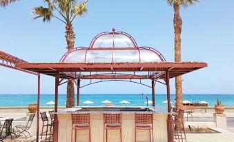 a beach bar with a red awning , surrounded by palm trees and chairs , under a clear blue sky at Grecotel Plaza Beach House