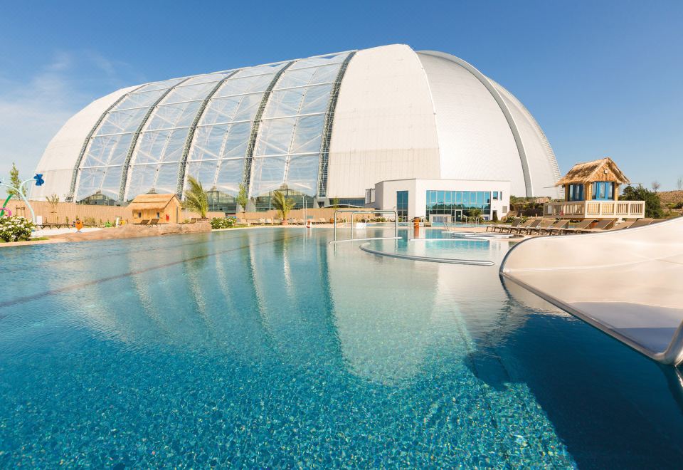 a large outdoor swimming pool surrounded by a building , with clear blue water and a white dome - shaped structure in the background at Tropical Islands