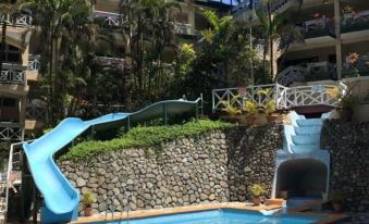 a swimming pool with a slide and jungle gym , surrounded by buildings and palm trees at Suva Motor Inn