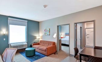 a modern living room with an orange couch , blue ottoman , and blue rug , connected to a bedroom with a bed and window at Home2 Suites by Hilton la Porte