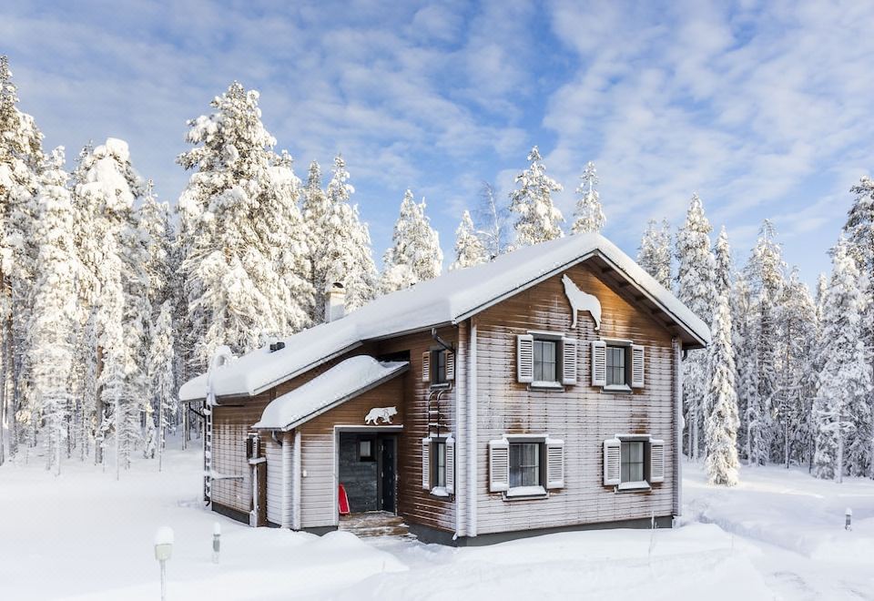 a wooden house surrounded by snow - covered trees , with a blue sky in the background at Ranua Resort Holiday Villas