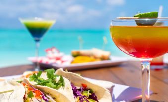 a beach scene with a table filled with food and drinks , including tacos , margaritas , and other drinks at Pelican Beach Hotel