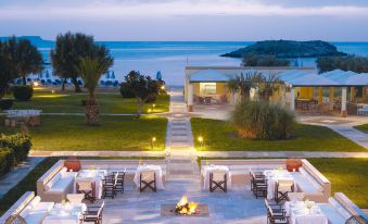 an outdoor dining area with a fire pit surrounded by chairs and tables , overlooking a body of water at Grecotel Meli Palace