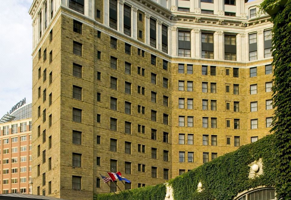 a tall , ornate hotel building surrounded by greenery and a grassy area , with a flag flying in front of it at The Saint Paul Hotel