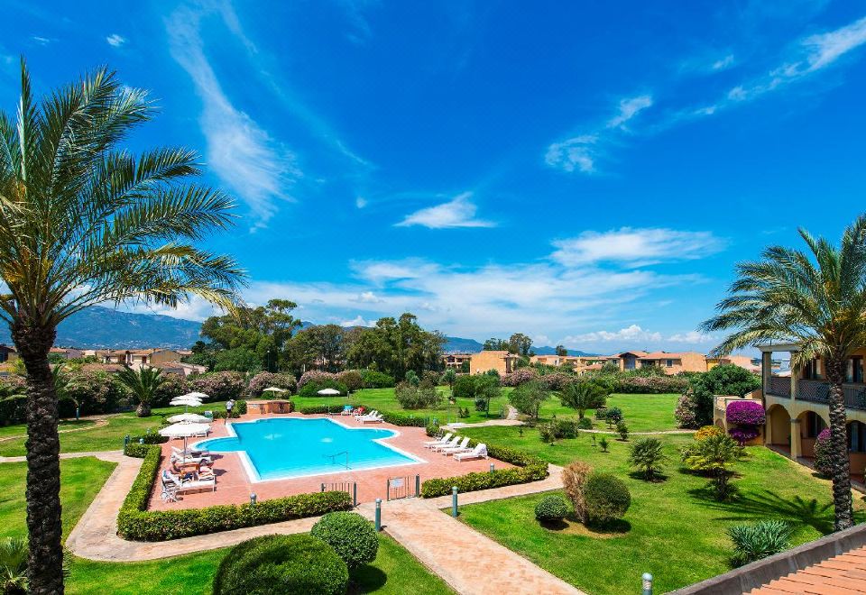 a large swimming pool is surrounded by a well - maintained lawn and palm trees , with a building in the background at Hotel Santa Gilla