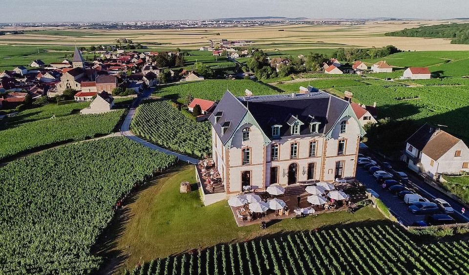 aerial view of a large white building surrounded by a vineyard , with several tables and chairs set up for an outdoor event at Chateau de Sacy