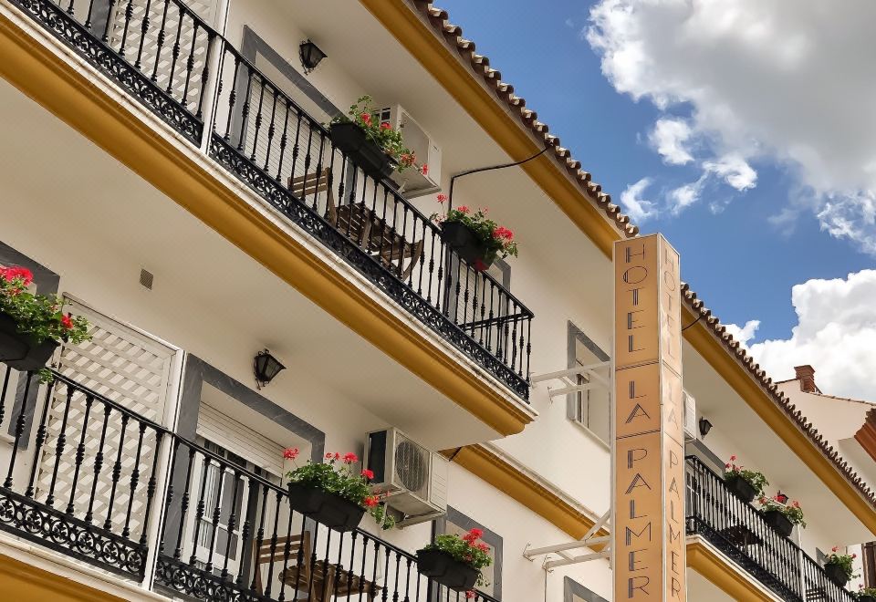 a white building with balconies adorned with potted plants , set against a blue sky dotted with clouds at La Palmera Rooms